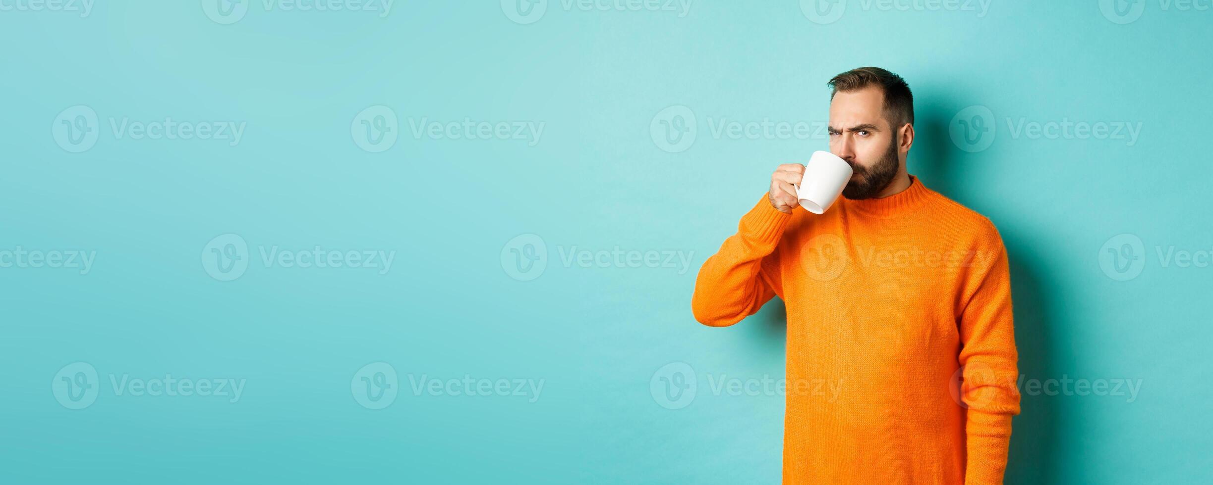Image of guy drinking coffee and looking suspicious at camera, stare at something strange, standing in orange sweater over turquoise background photo