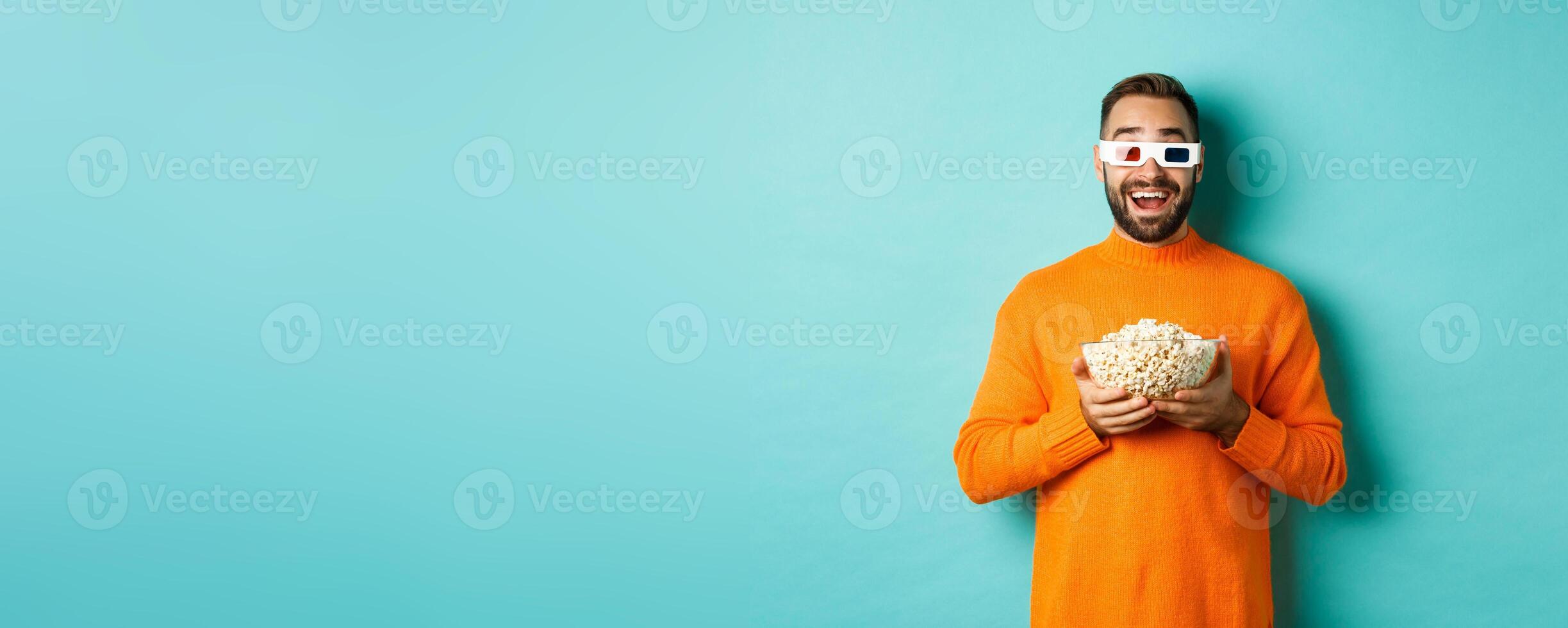 Happy guy watching movies in 3d glasses, eating popcorn and looking at camera, standing over light blue background photo