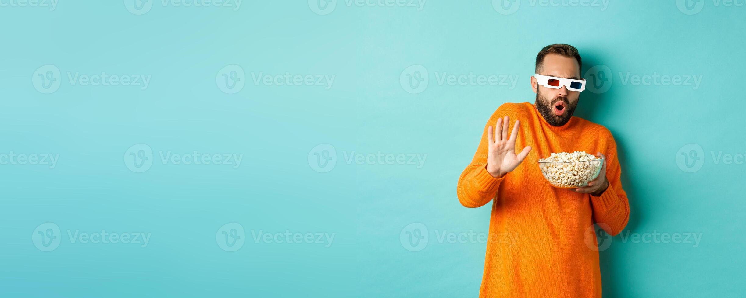 Image of man in 3d glasses watching movie, getting scared of special effects, looking in awe, standing with popcorn against white background photo