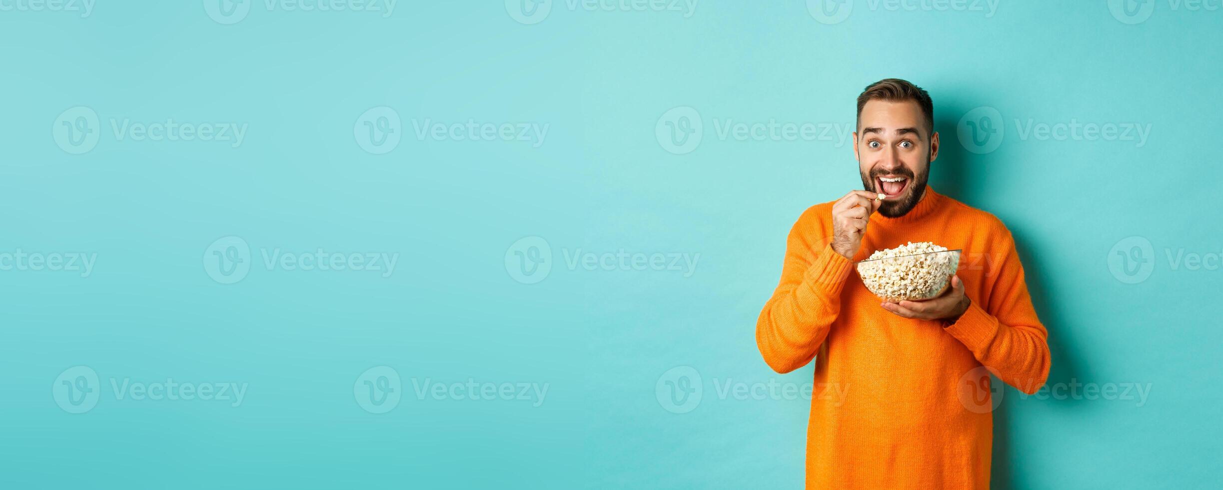 Excited young man watching interesting movie on tv screen, eating popcorn and looking amazed, blue background photo