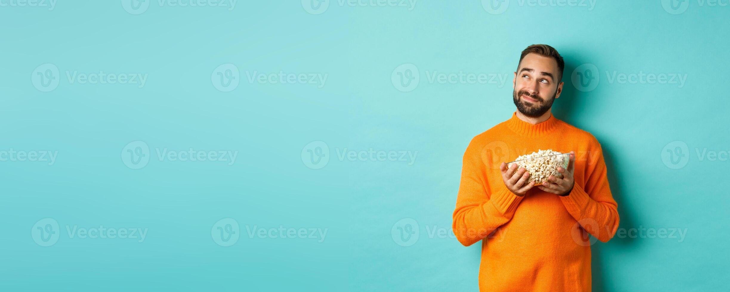 Handsome young man in orange sweater, looking thoughtful at upper left corner, holding popcorn, picking movie, blue background photo