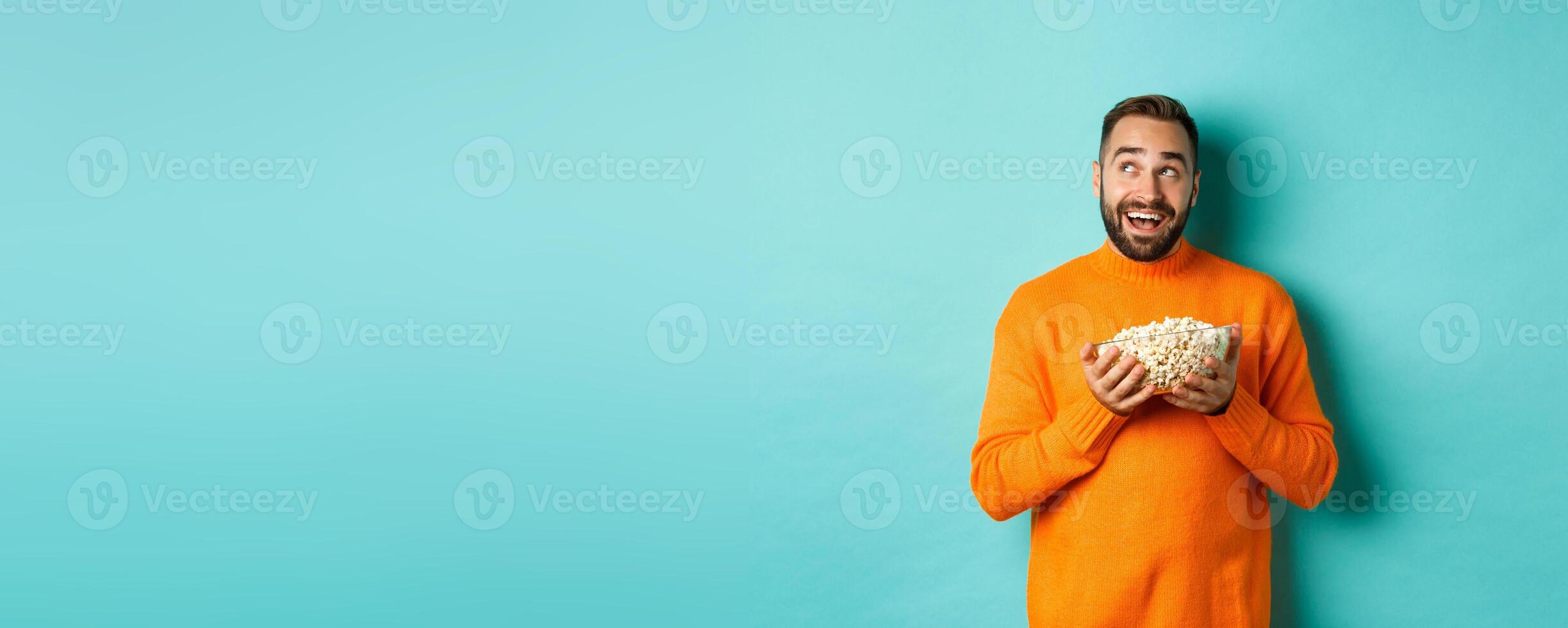 Handsome young man in orange sweater, looking thoughtful at upper left corner, holding popcorn, picking movie, blue background photo