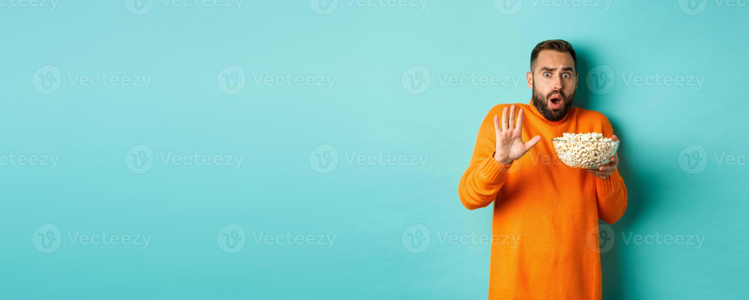 Portrait of scared guy holding bowl of popcorn and declining something, showing stop gesture, standing over blue background photo