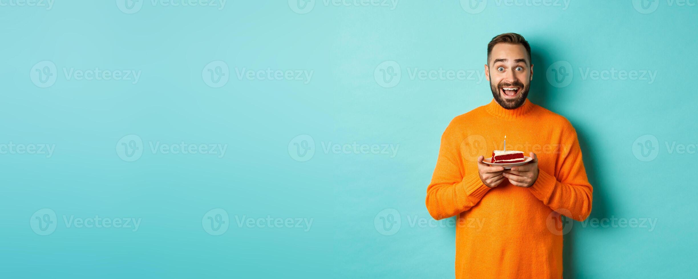 Happy adult man celebrating birthday, holding bday cake with candle and smiling, standing against turquoise background photo
