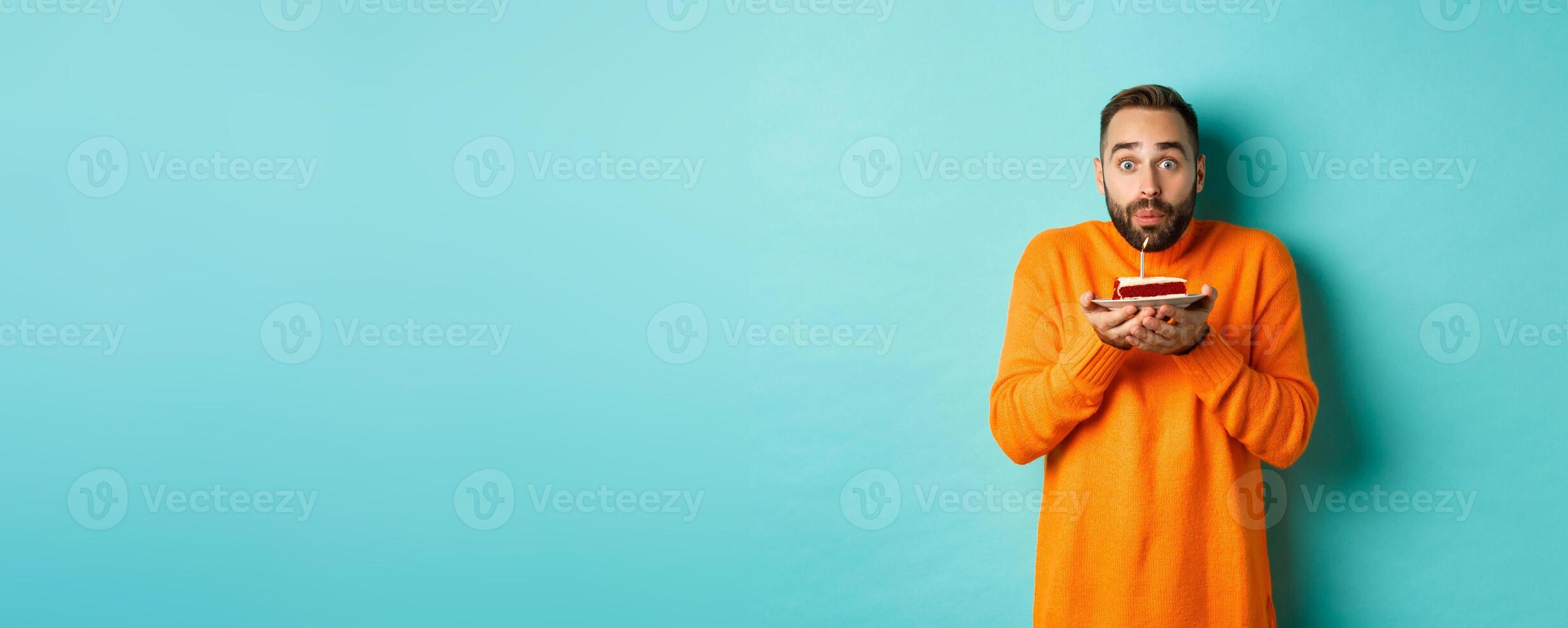 Handsome adult man celebrating birthday, blowing out candle on cake and making wish, standing against turquoise background photo