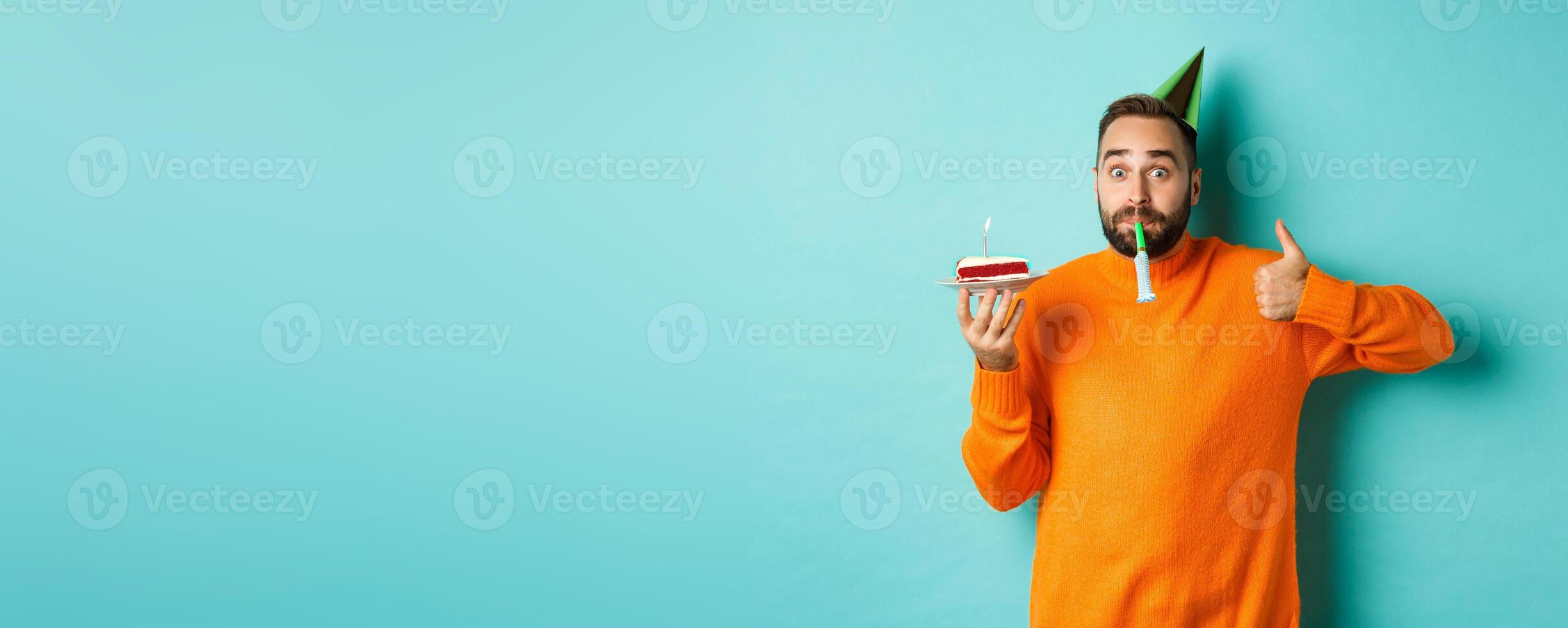 Happy birthday guy celebrating, wearing party hat, blowing wistle and holding bday cake and showing thumb-up in approval, standing over turquoise background photo