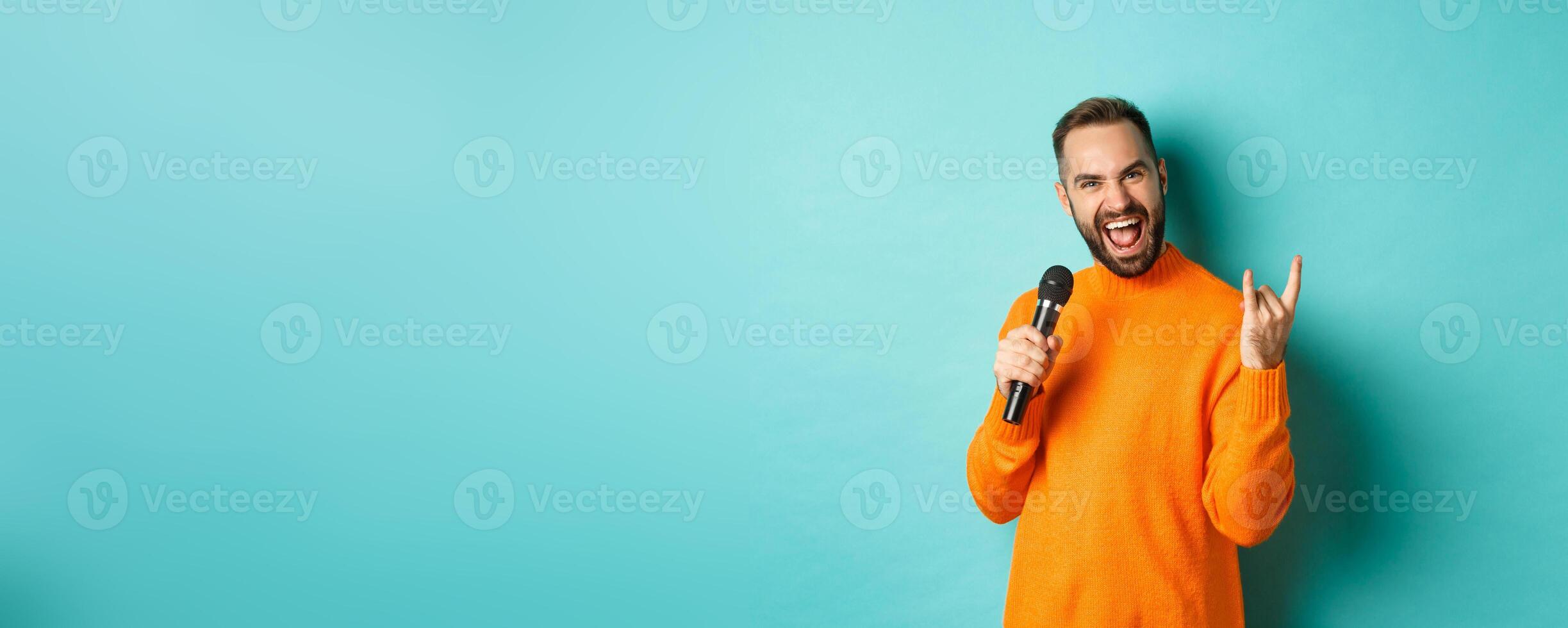 Joyful handsome man singing song in microphone, showing rock-n-roll gesture excited, standing over turquoise background photo