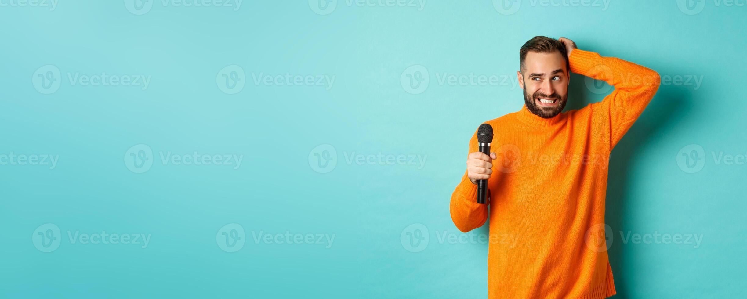 Shy and awkward adult man scratching head, holding microphone before singing, standing over blue background photo