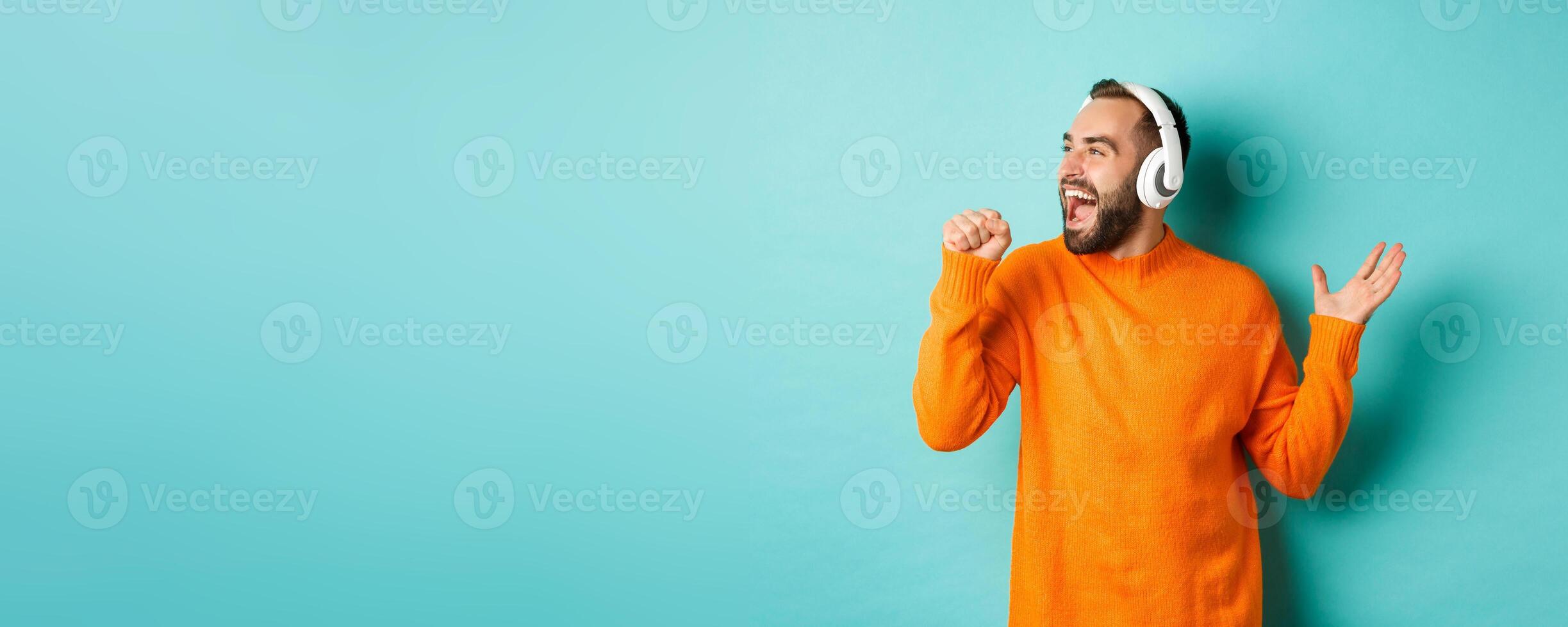 Happy man lipsync while listening music in headphones, holding invisible microphone, standing over turquoise background photo