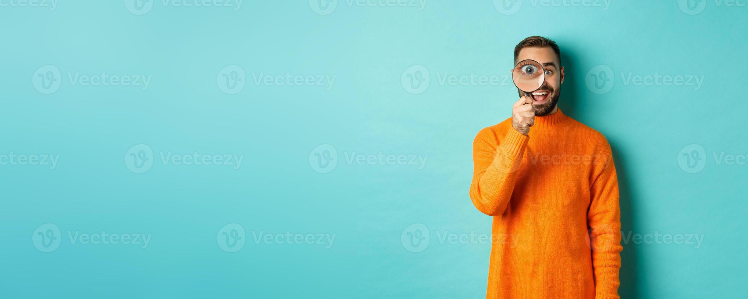 Cheerful man searching for something, looking through magnifying glass and smiling happy, standing over blue background photo