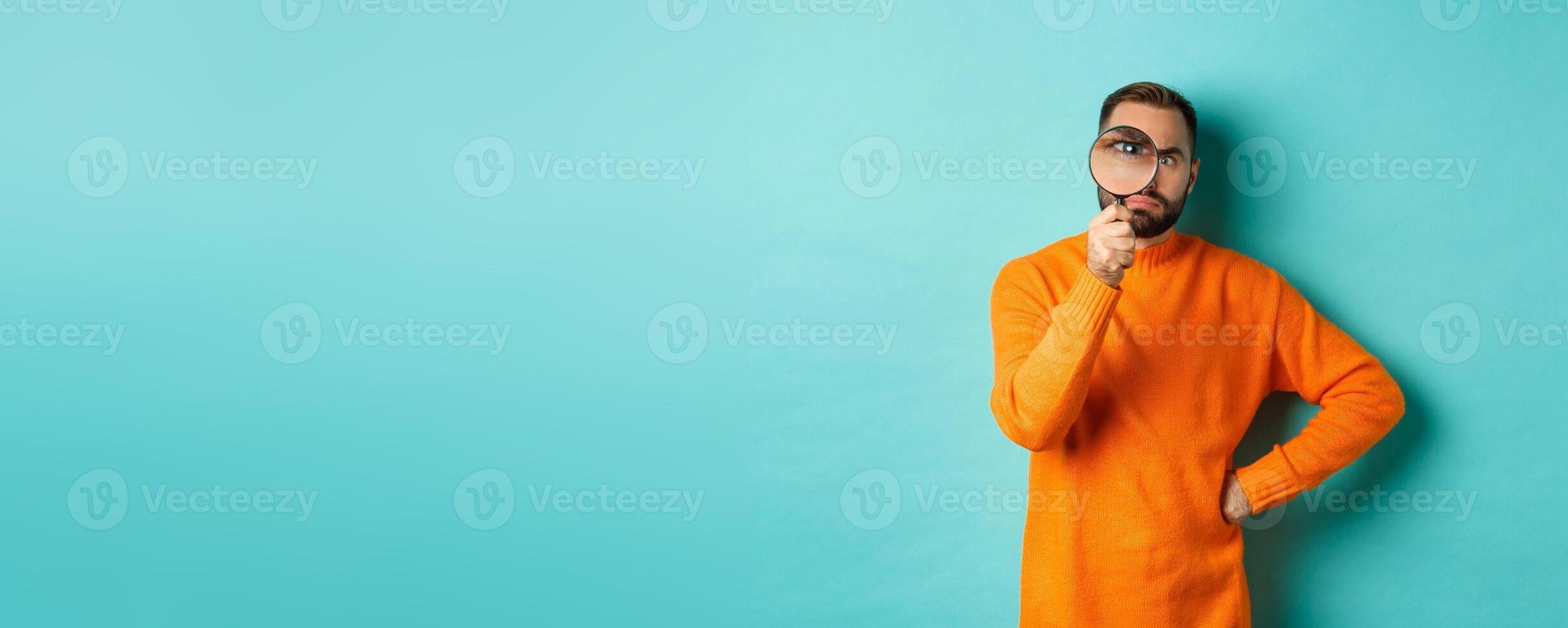 Funny man looking serious through magnifying glass, inspecting something, standing in orange sweater against turquoise background photo