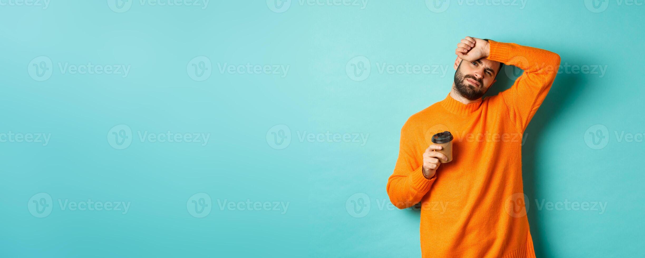 Tired adult man taking break from work and drinking coffee, wipe sweat off forehead and looking exhausted, standing in orange sweater over turquoise background photo