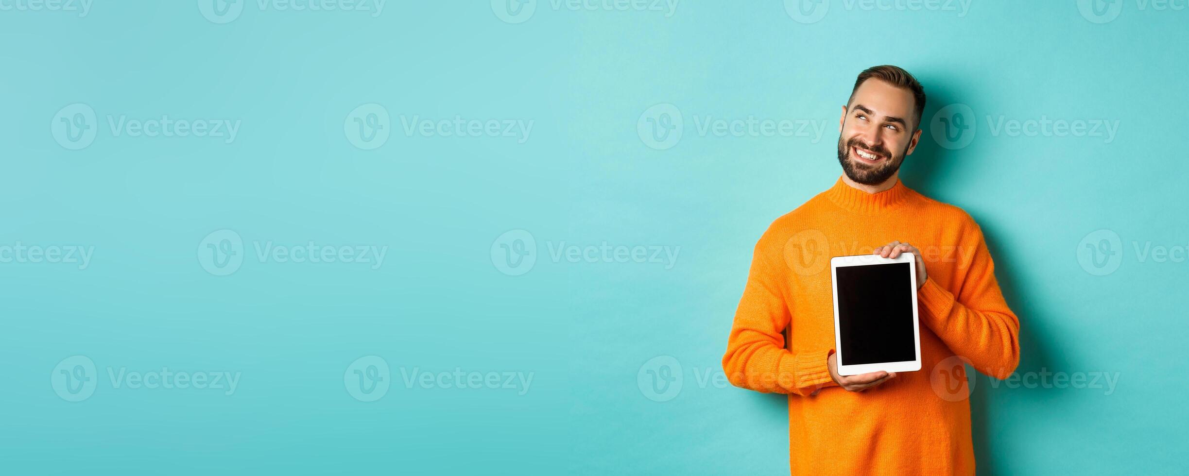 Technology. Handsome adult man showing digital tablet screen, smiling and looking at upper left corner thoughtful, imaging or daydreaming, blue background photo