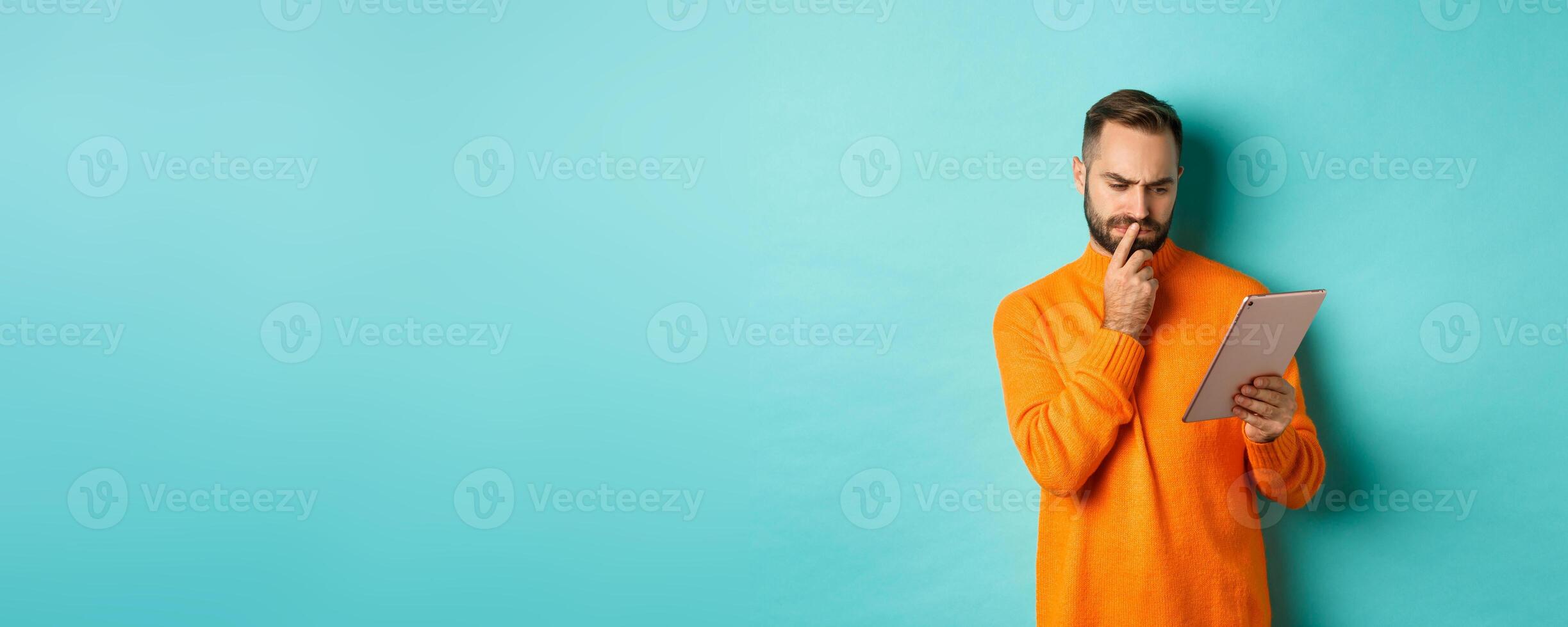 Serious man pondering and looking at digital tablet screen, reading social media, standing over turquoise background photo