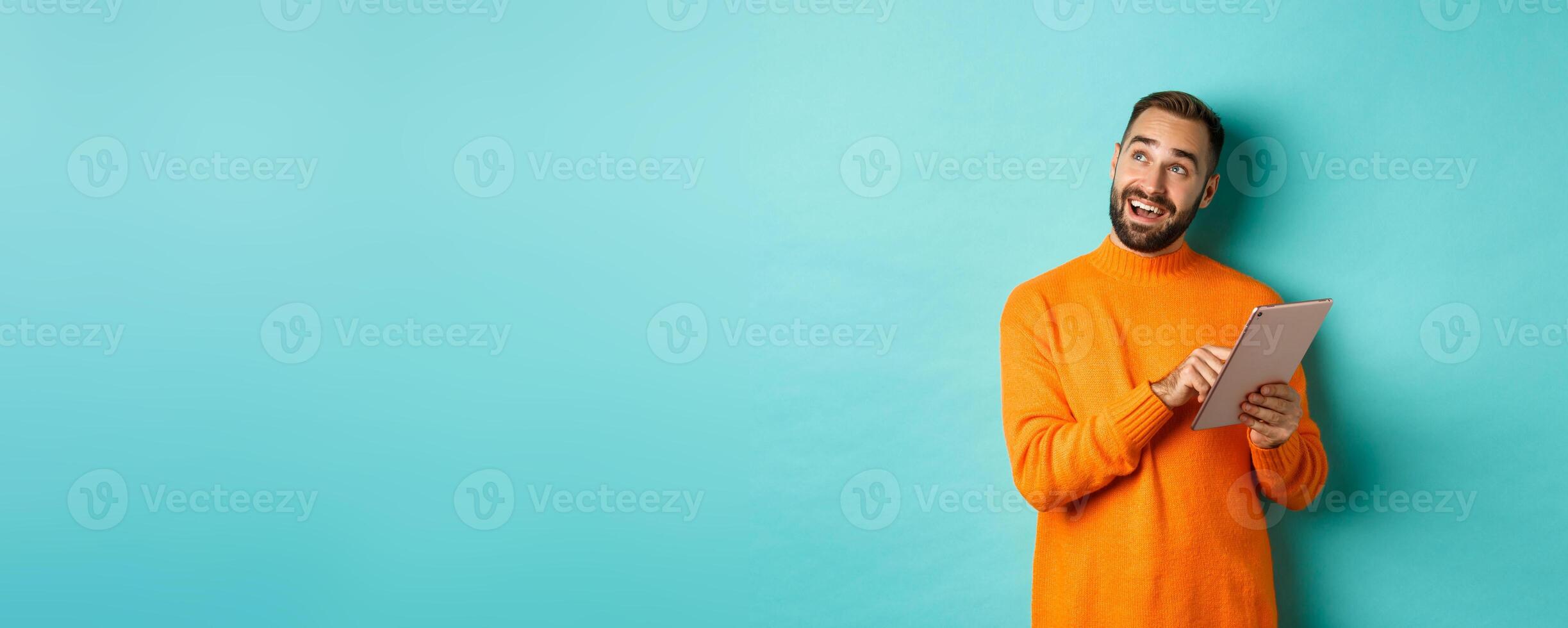 Handsome man thinking and using digital tablet, shopping online and pondering ideas, looking at upper left corner, standing over light blue background photo