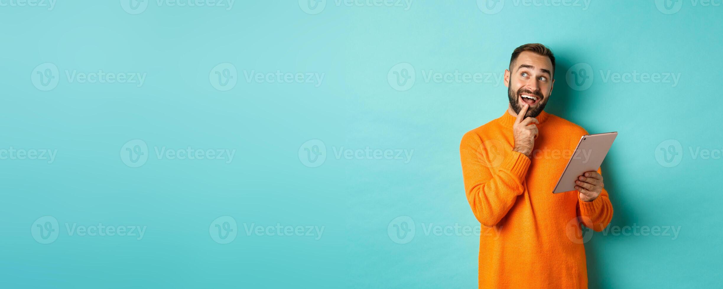 Handsome man thinking and using digital tablet, shopping online and pondering ideas, looking at upper left corner, standing over light blue background photo