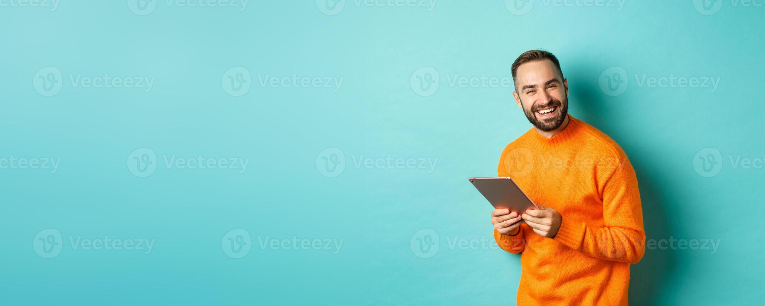 Handsome bearded man using digital tablet, laughing at camera, standing happy against light blue background photo