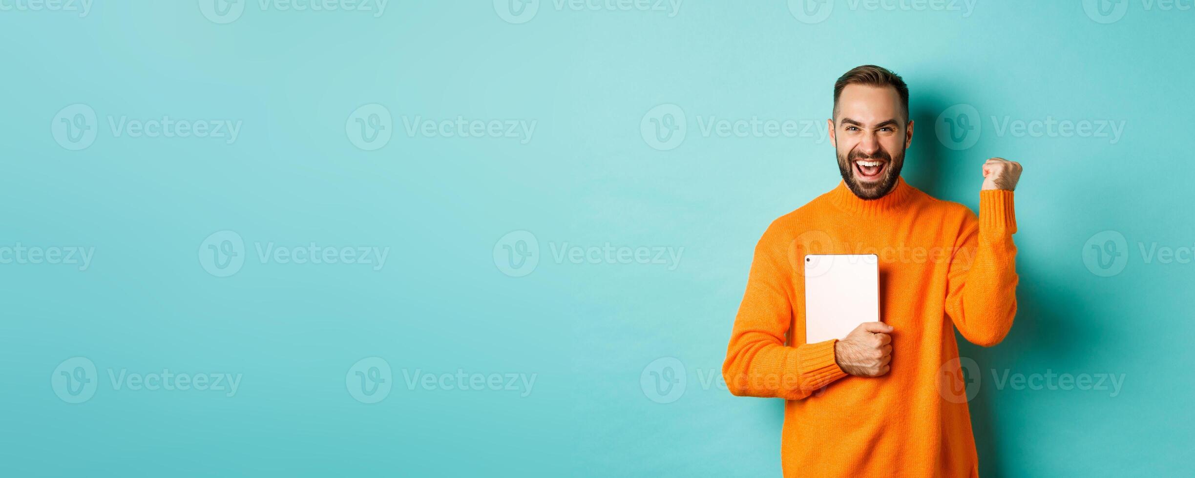 Excited adult man holding laptop and making fist pump, rejoicing of win, standing satisfied against turquoise background photo