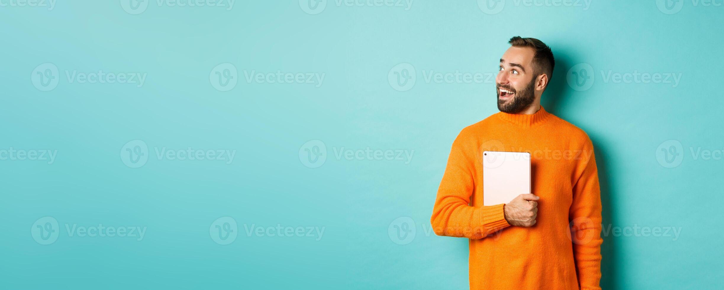 Handsome man holding laptop, looking left with surprise and amazement, standing in orange sweater against light blue background photo