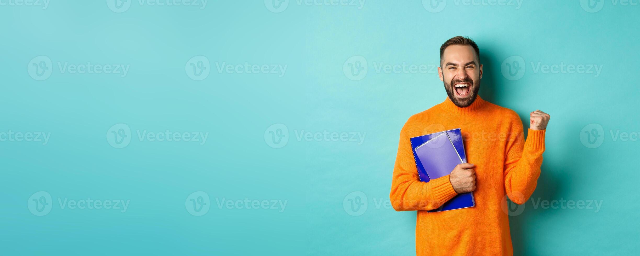 Education. Happy and satisfied man rejoicing, holding notebooks and making fist pump, shouting for joy, standing against turquoise background photo
