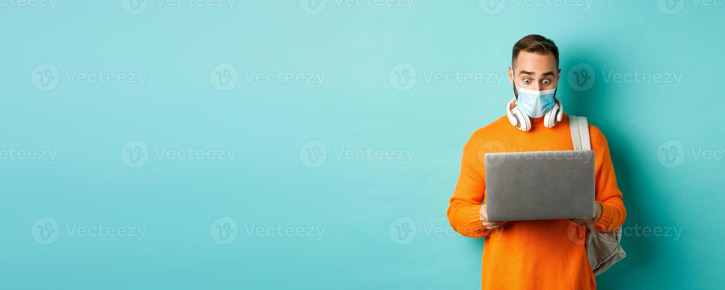 Happy young man in medical mask, working on laptop, looking shocked at computer screen, standing over light blue background photo
