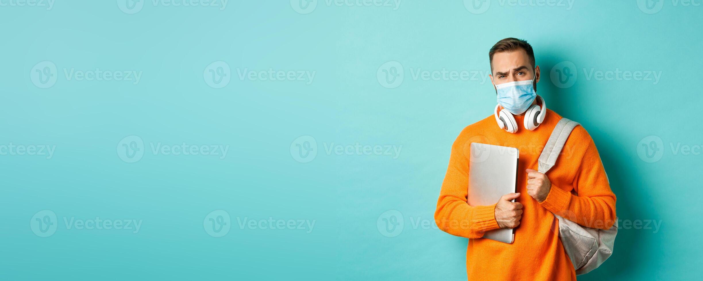 Handsome caucasian man with headphones and backpack, holding laptop and wearing medical mask, looking doubtful and skeptical, standing over light blue background photo