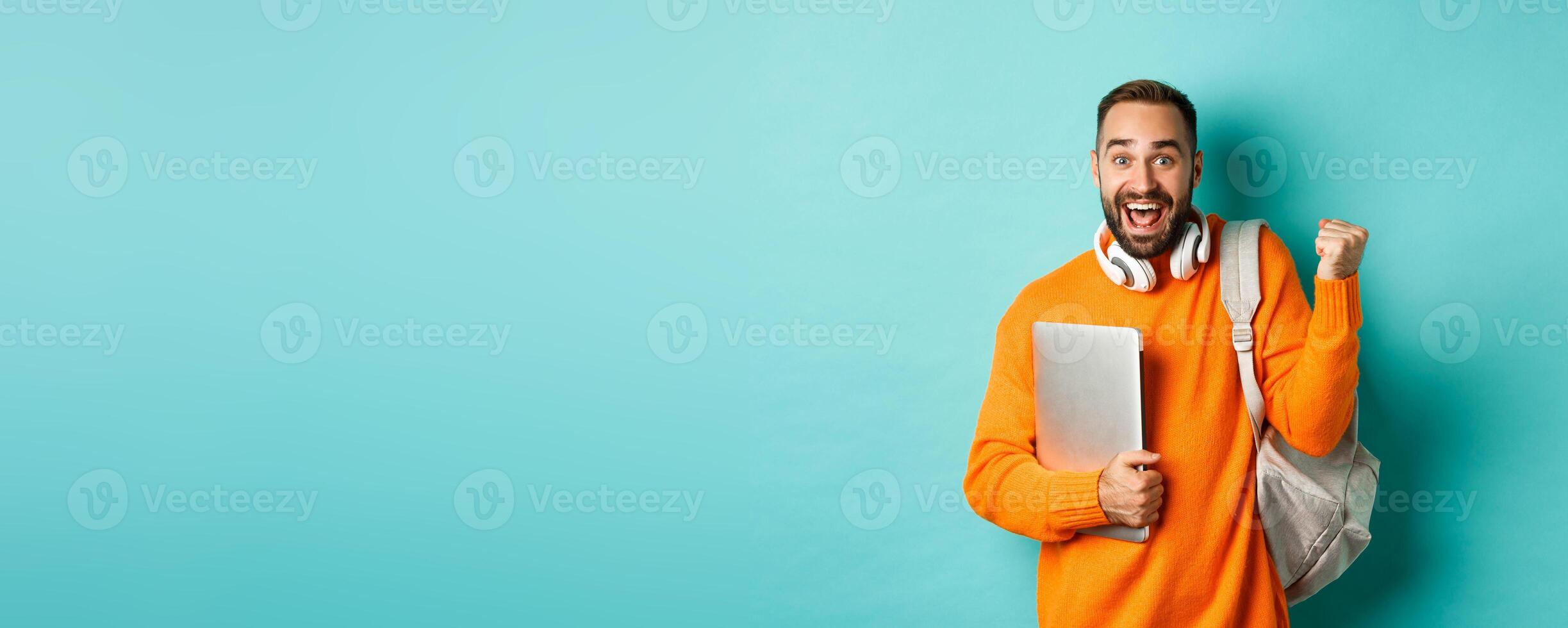 Happy man with backpack and headphones, holding laptop and smiling, cheering of win, triumphing over turquoise background photo
