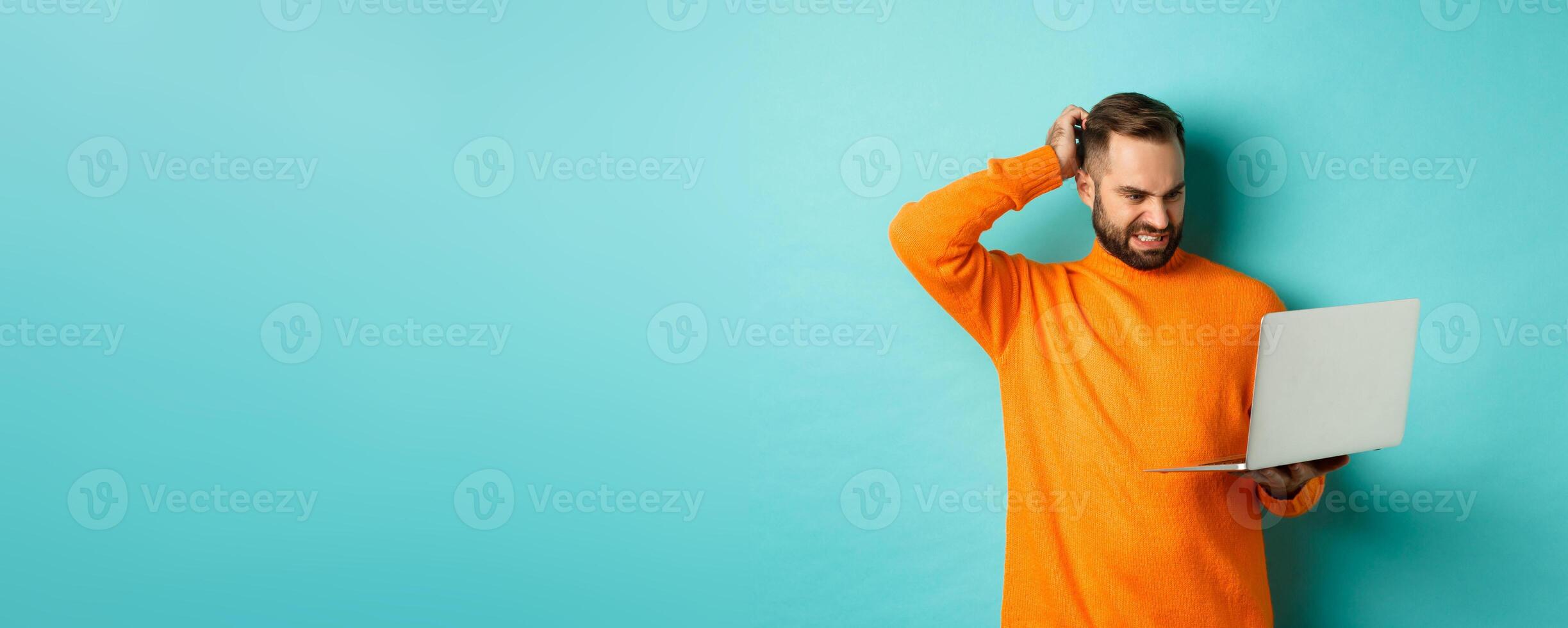 Freelance and technology concept. Confused man scratching head and looking at laptop screen puzzled, dont understand, standing over light blue background photo