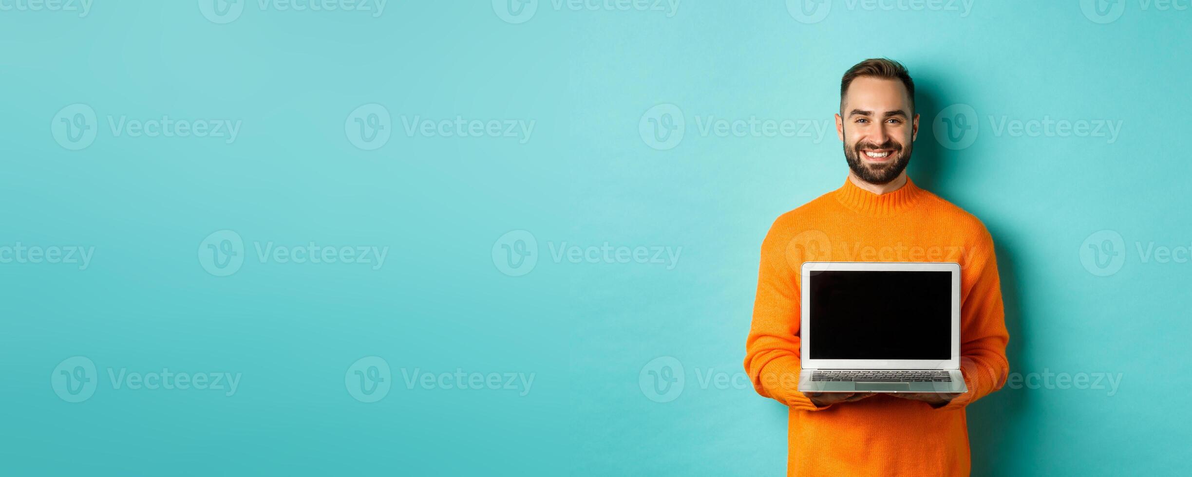 Handsome bearded man in orange sweater showing laptop screen, demonstrating online store, standing over light blue background photo