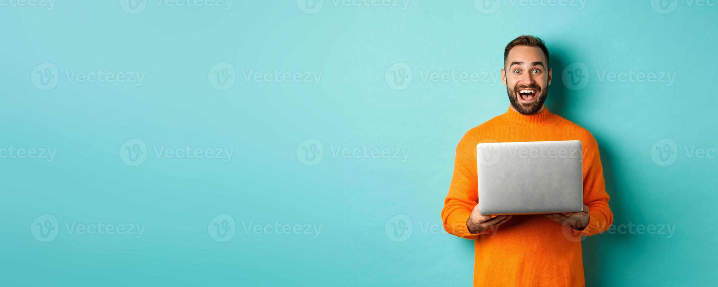 Happy man using laptop and looking excited at camera, standing with computer against light blue background photo