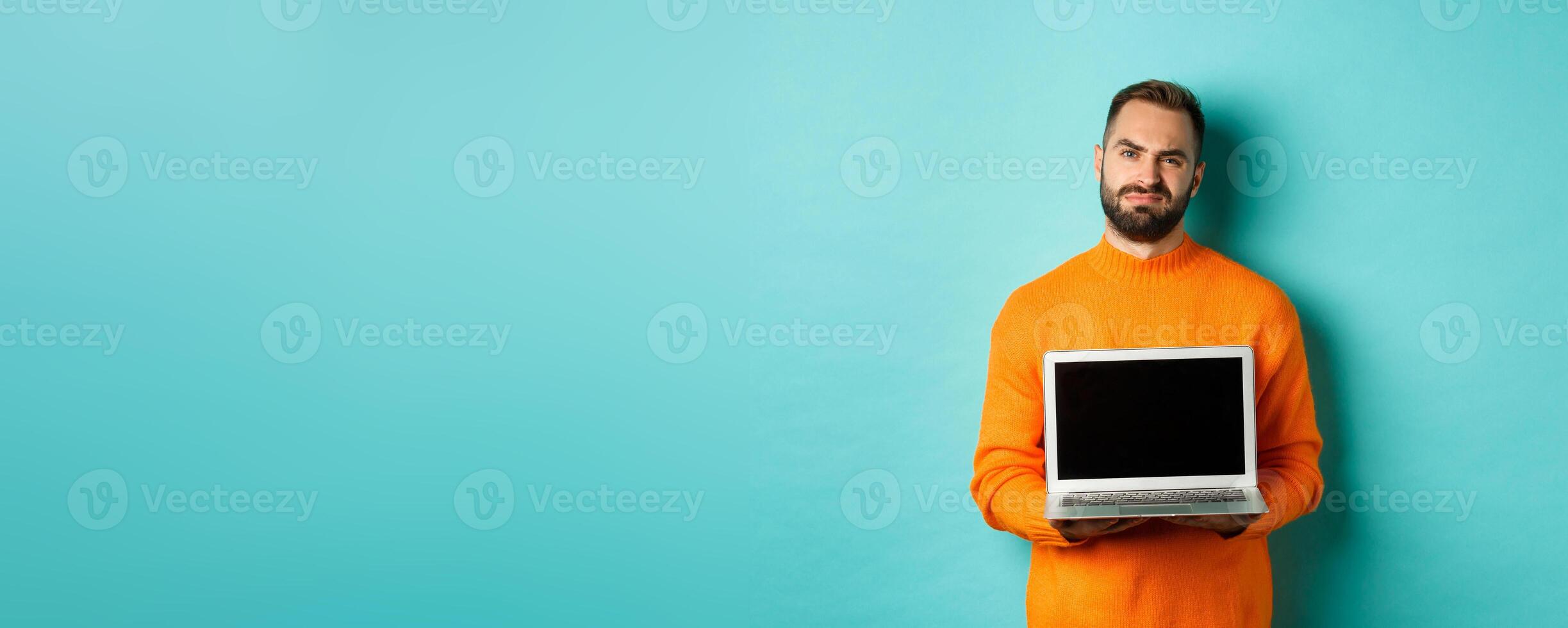 Handsome bearded man in orange sweater showing laptop screen, demonstrating promo, grimacing disappointed and upset standing over light blue background photo
