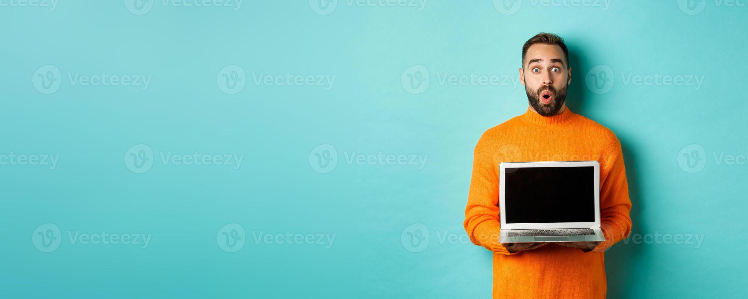 Handsome bearded man in orange sweater showing laptop screen, demonstrating promo, standing over light blue background photo