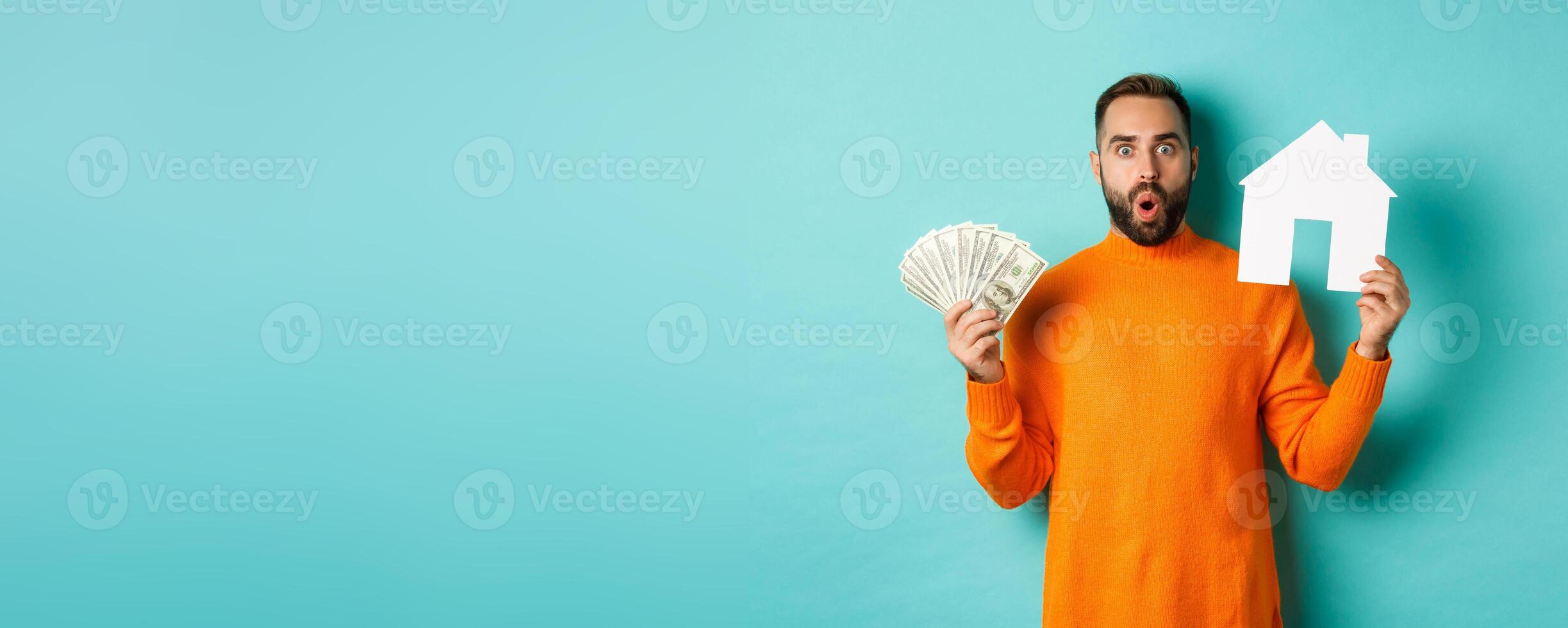 Real estate concept and mortgage concept. Amazed buyer holding money and paper house maket, buying apartment, standing over light blue background photo