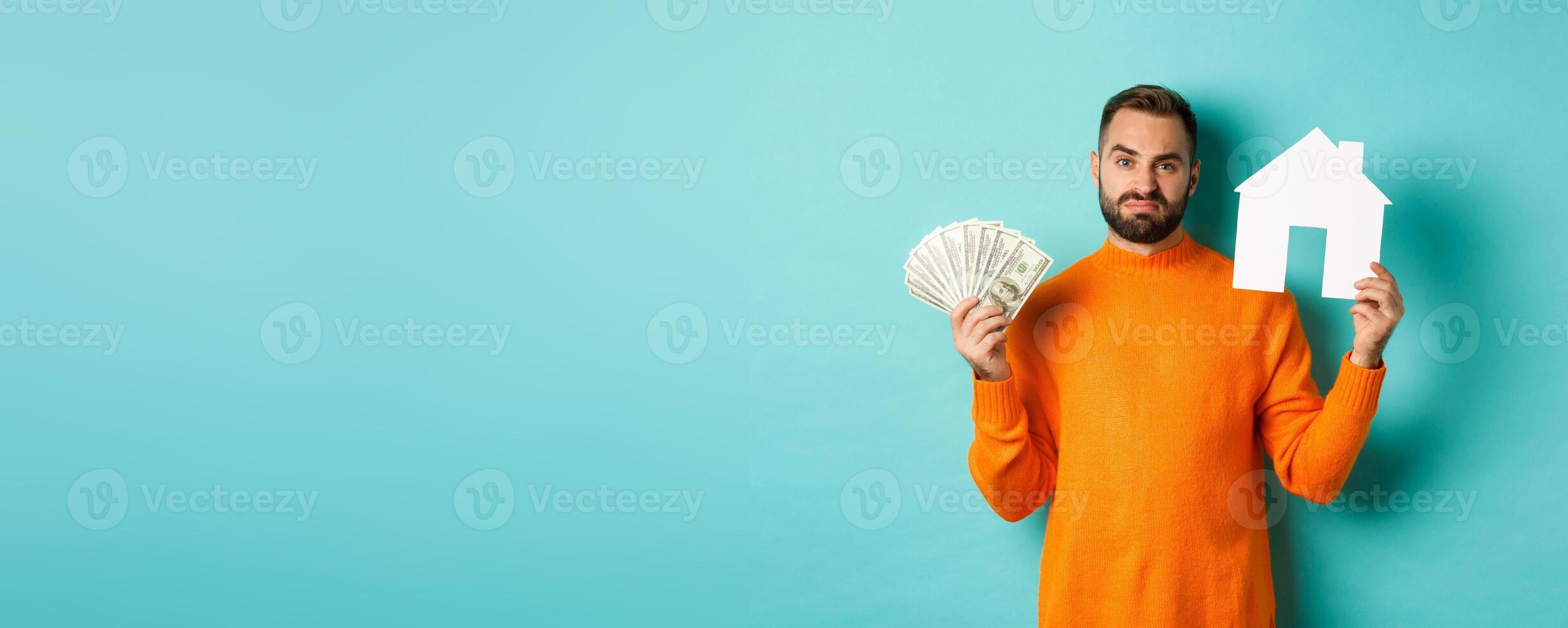 Real estate concept and mortgage concept. Sad man showing house maket and money, looking upset and disappointed, standing over light blue background photo
