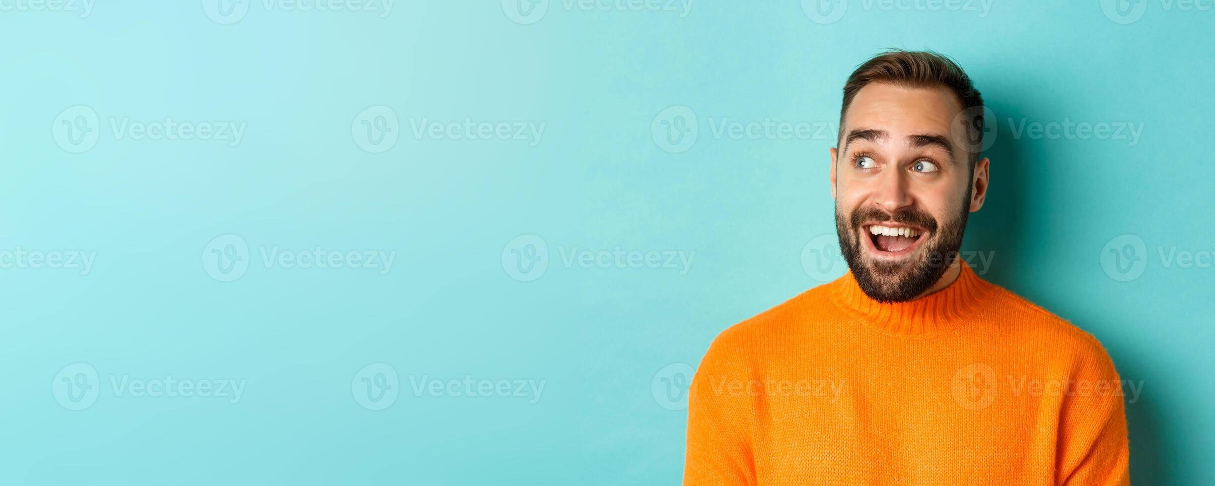 Close-up of handsome caucasian man smiling, looking left with surprised face, staring at logo, wearing orange sweater, standing against turquoise background photo