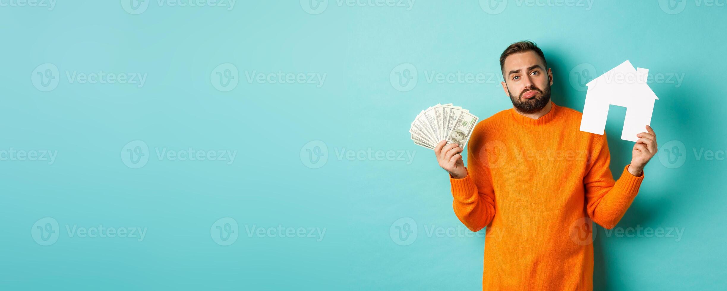 Real estate concept and mortgage concept. Sad and confused man showing money and paper house maket, shrugging upset, standing over light blue background photo
