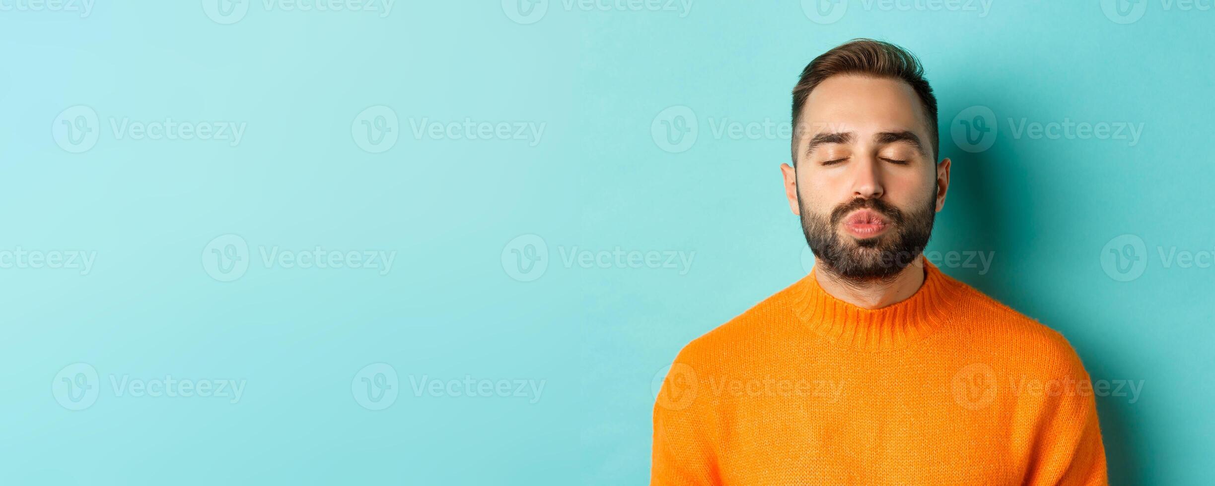 Close-up of young man in sweater pucker lips, close eyes and waiting for kiss, standing over light blue background photo