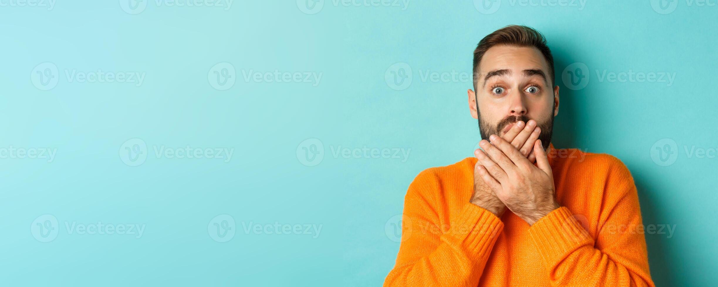 Close-up of shocked caucasian man covering his lips and mouth, staring astounded at camera, standing against turquoise background photo