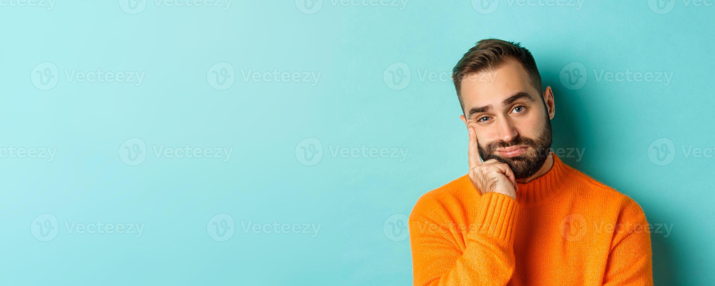 Close-up of unamused sad man looking at camera, pouting and leaning face on hand, standing over light blue background photo