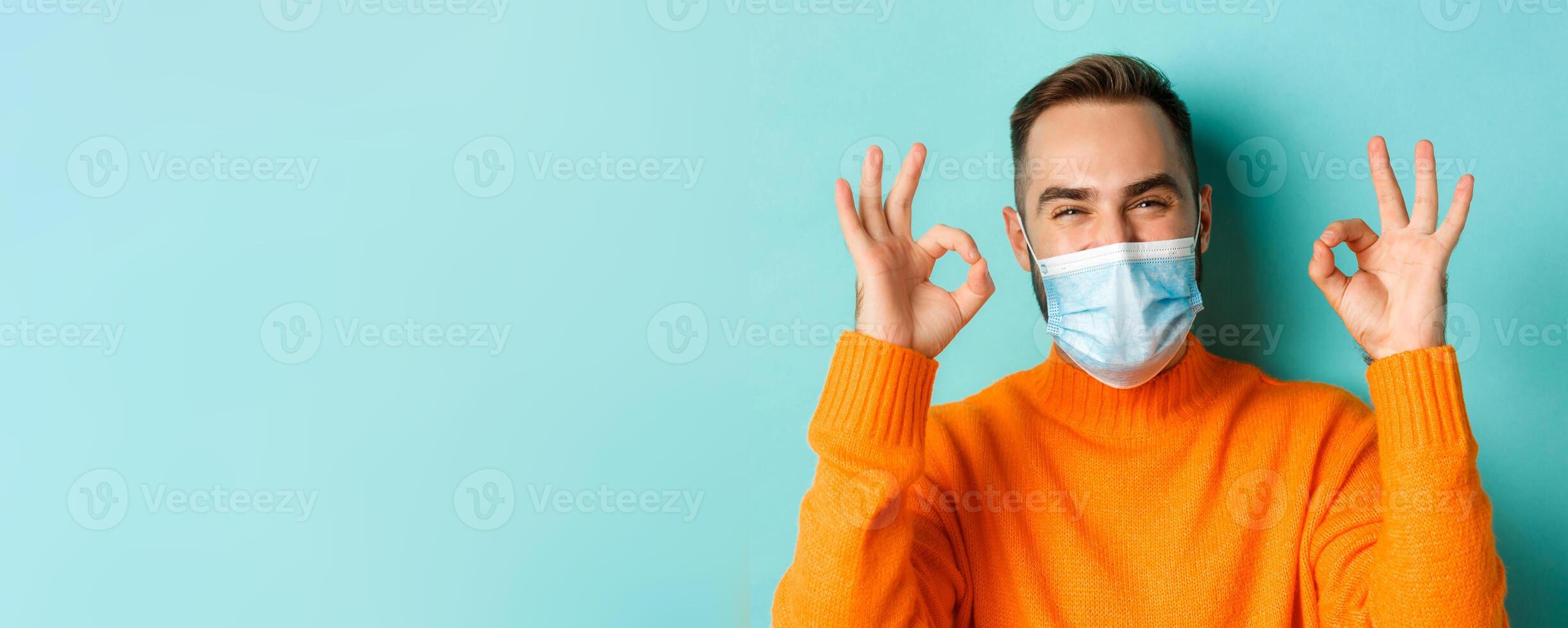Covid-19, social distancing and quarantine concept. Close-up of satisfied male model in medical mask showing okay signs, praising and agreeing, standing over light blue background photo