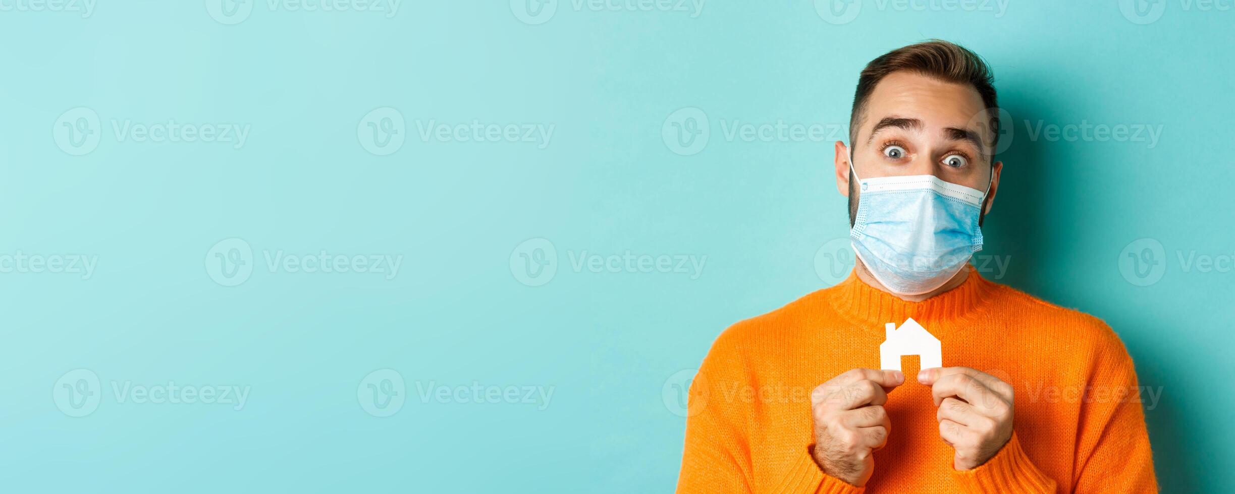Real estate and coronavirus pandemic concept. Close-up of adult man in medical mask holding small paper house maket and smiling, searching for apartment, light blue background photo