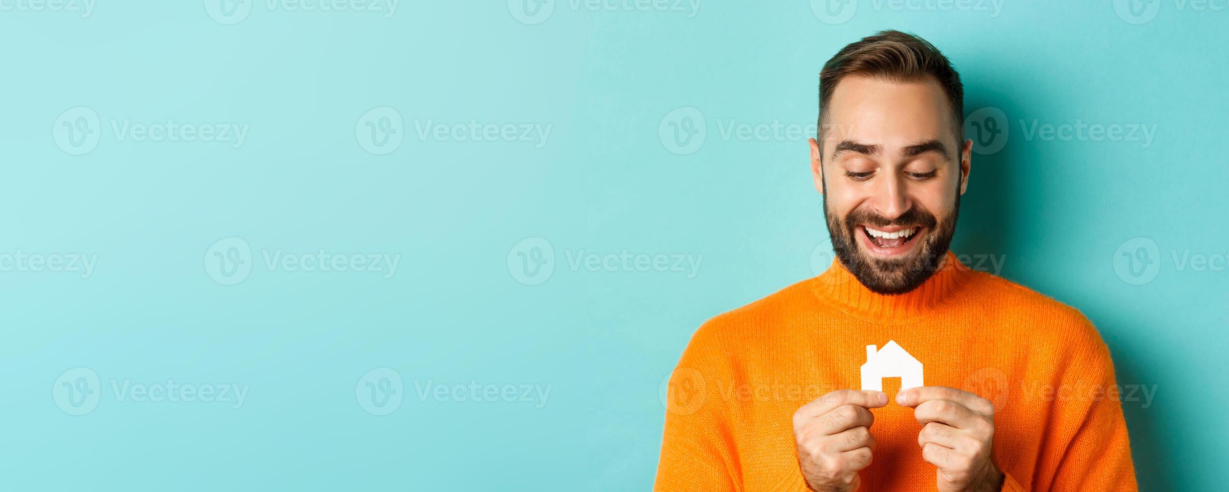 Real estate concept. Happy young man searching for home rent, holding house paper maket and smiling, standing over blue background photo