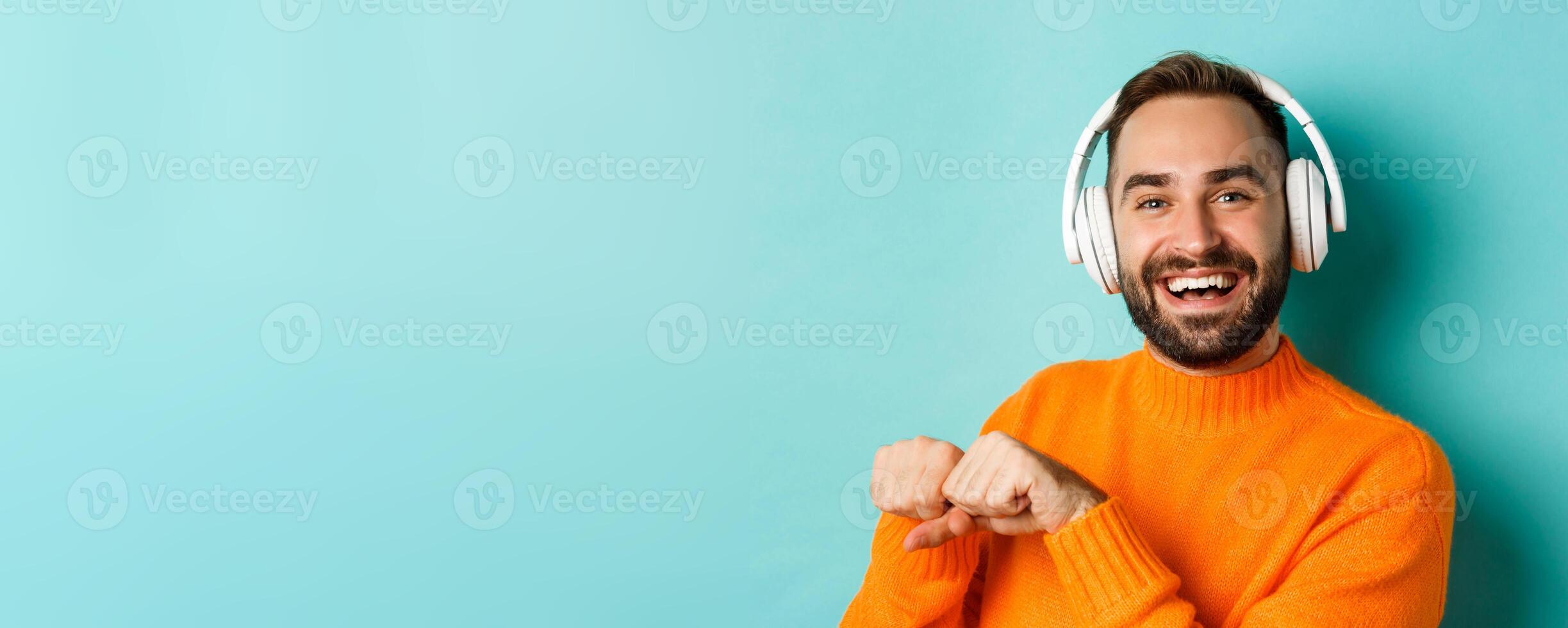 Close-up of handsome modern man listening music in headphones, standing in orange sweater over turquoise background photo