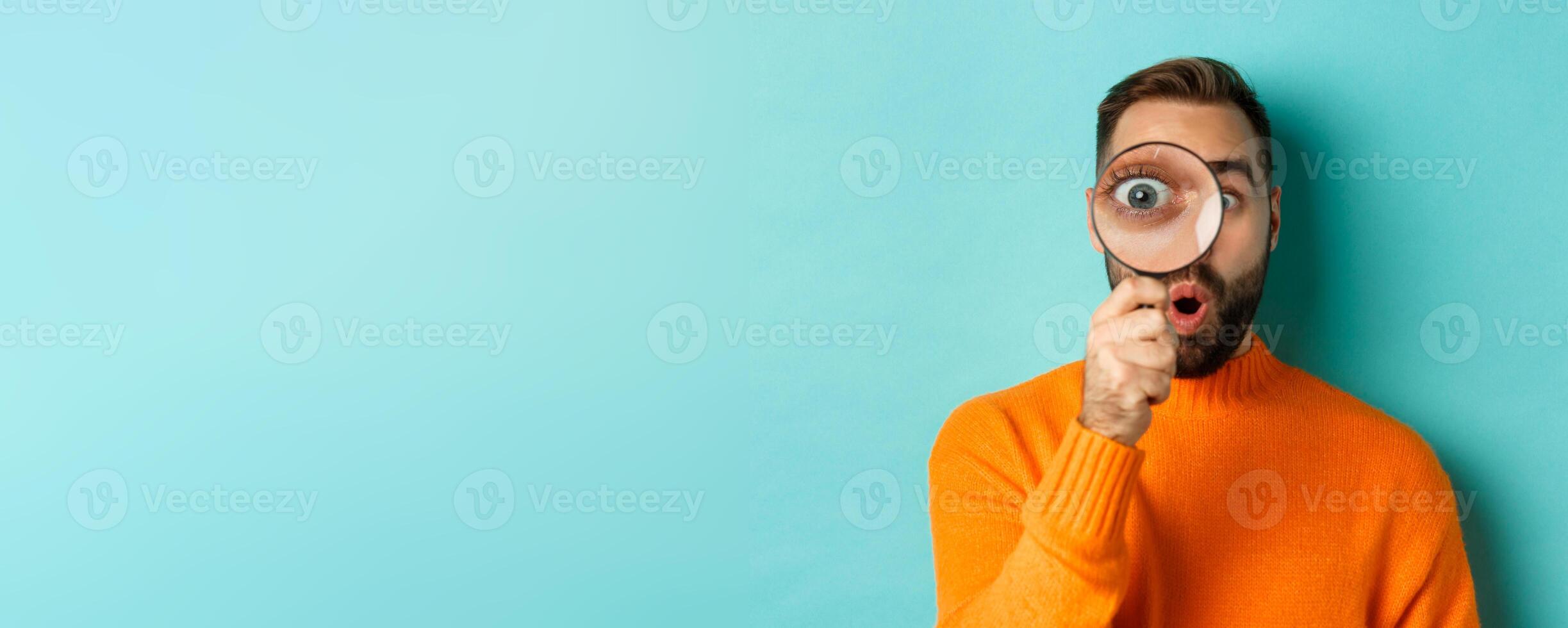 Funny man looking through magnifying glass, searching or investigating something, standing in orange sweater against turquoise background photo