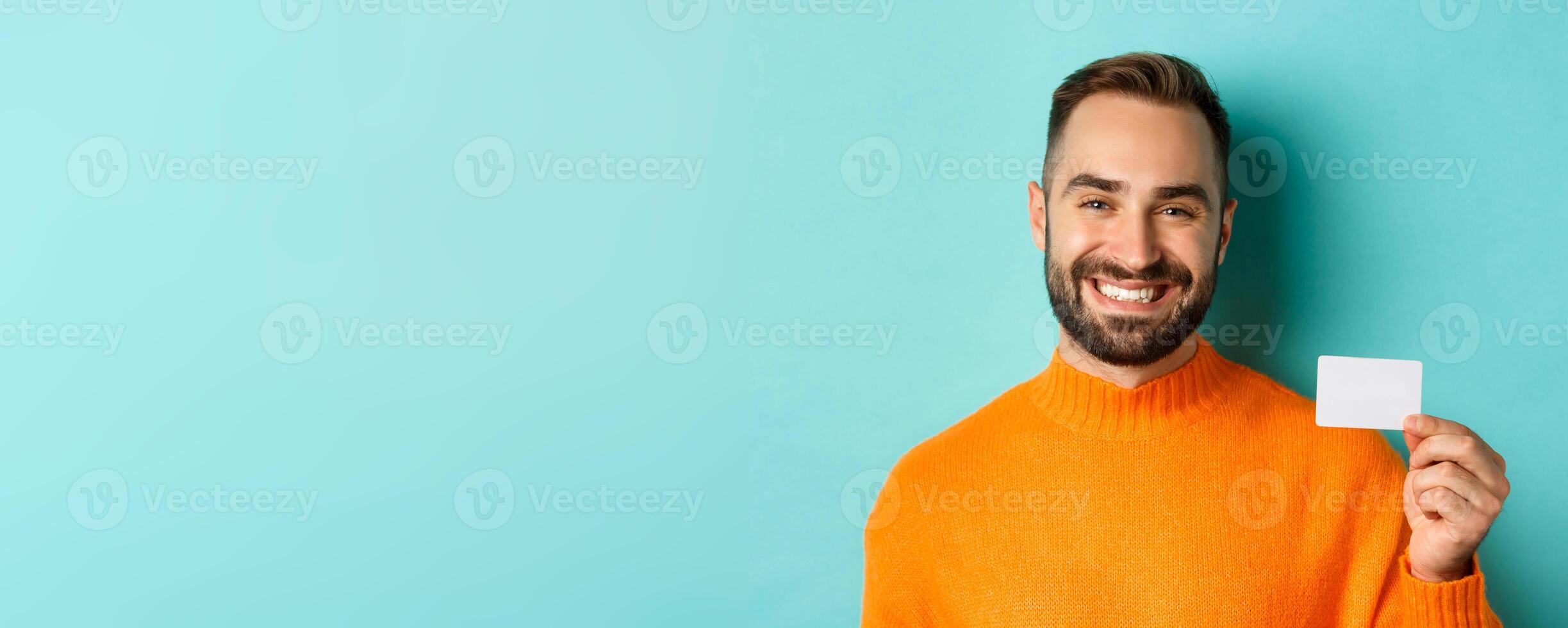 Close-up of handsome caucasian man going on shopping, showing credit card and smiling, standing over turquoise background photo