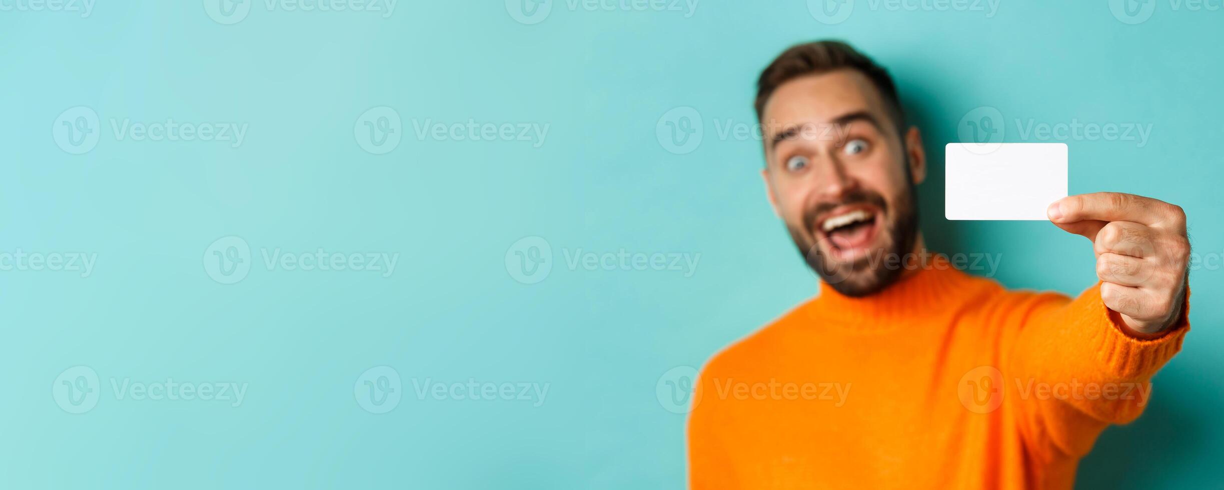 Close-up of handsome caucasian man going on shopping, showing credit card and smiling, standing over turquoise background photo