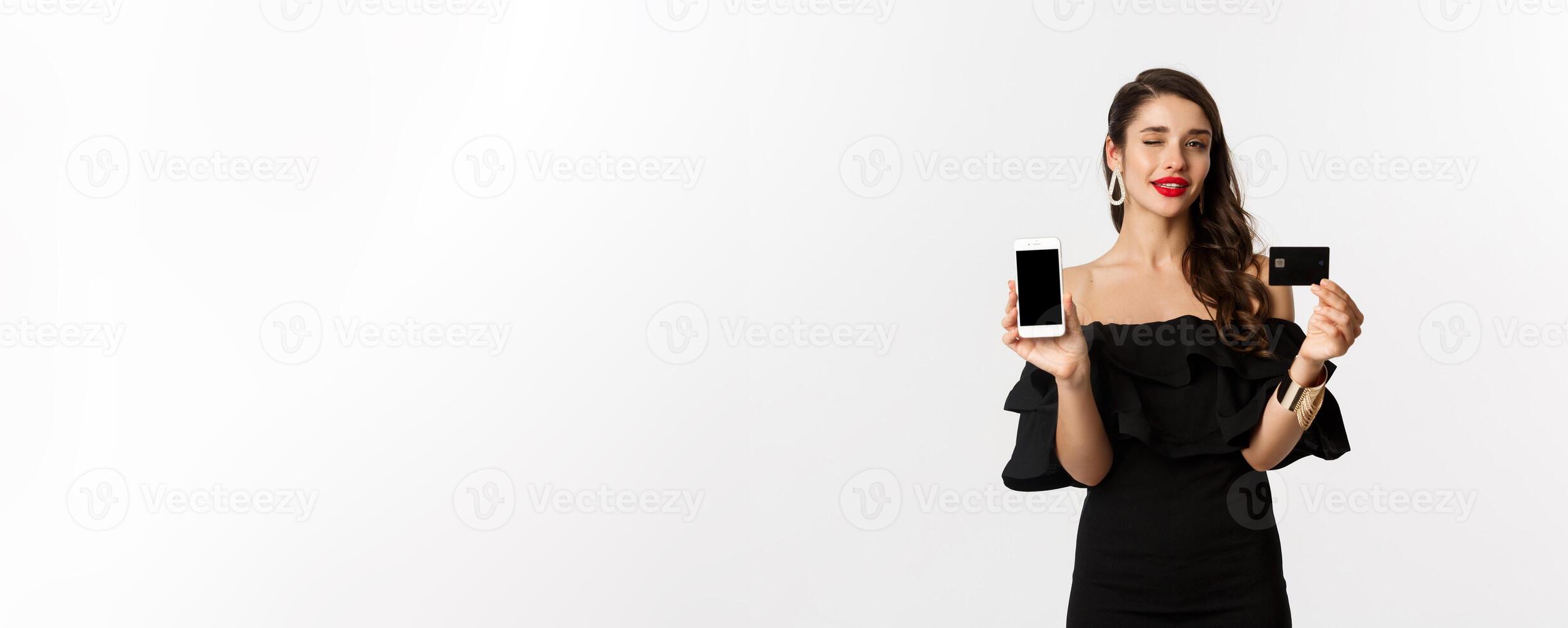 Fashion and shopping concept. Beautiful woman with red lips, winking at camera, showing smartphone screen and credit card, buying online, white background photo