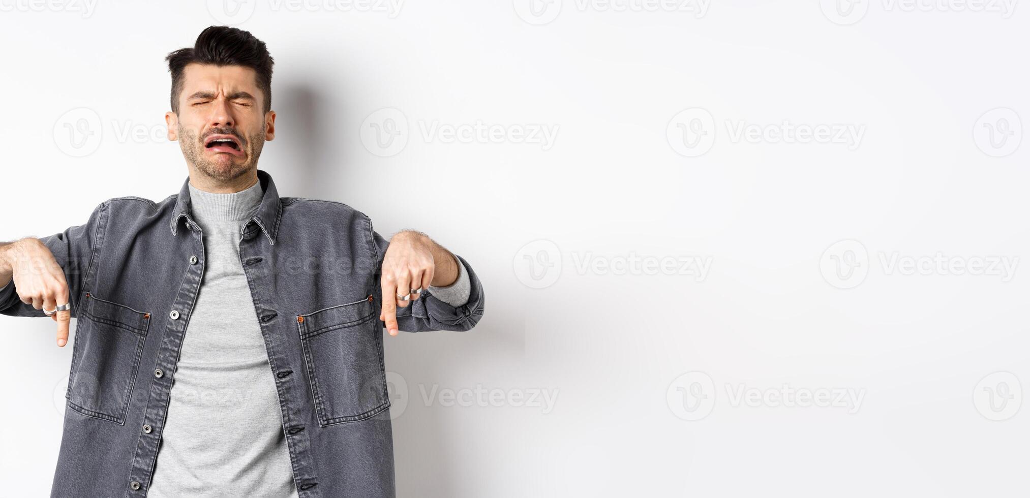 Sad crying man pointing fingers down, sobbing miserable, showing upsetting news, standing in denim jacket on white background photo