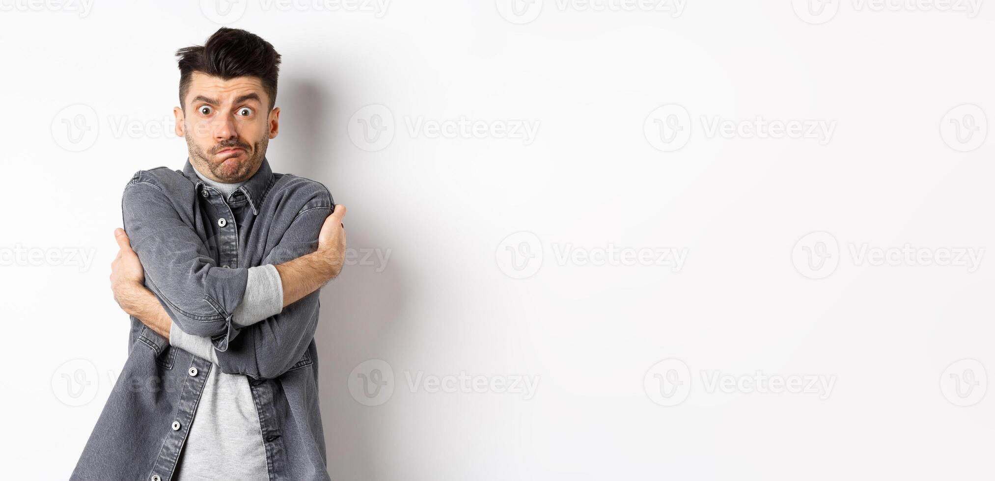 Funny young man hugging himself and grimacing, feeling embarrassed and exposed, covering body from people glances, white background photo