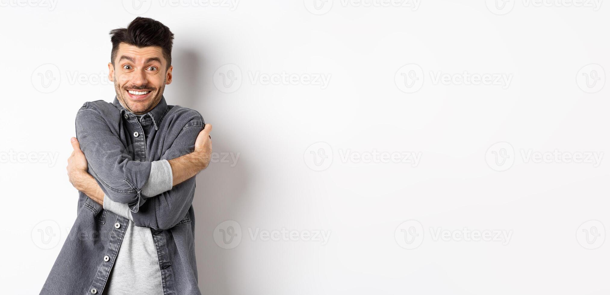 Happy handsome man hugging himself and smiling, embracing own body, standing on white background in grey jacket photo